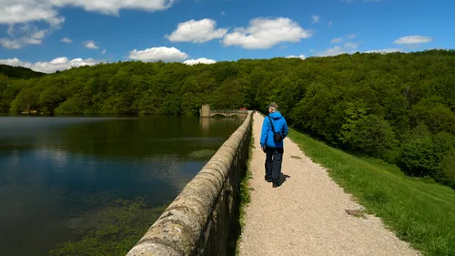 This landscape photograph captures a scene at Linacre reservoir in Derbyshire, England, United Kingdom, during the early afternoon in the spring season. The image features a walker, identified as Michael Weinbren, dressed in a blue jacket, strolling along the dam that borders the tranquil water of the lake. Lush green woodland stretches across the background, highlighting the vibrant spring foliage characteristic of Linacre. The dam structure is visible in the distance, and the peaceful water reflects the clear blue sky and scattered clouds, creating a picturesque setting. This nature photograph beautifully showcases the scenic beauty and serene atmosphere of Linacre reservoir, registering the essence of Derbyshire's landscape in England.